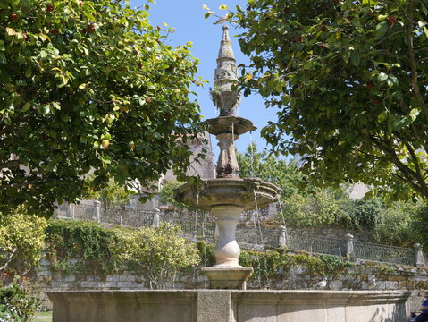 View of the historic stone fountain in Fonte Plaza de Orense, framed by lush green trees and stone walls in the city center, Pontevedra, Pontevedra, Spain.
