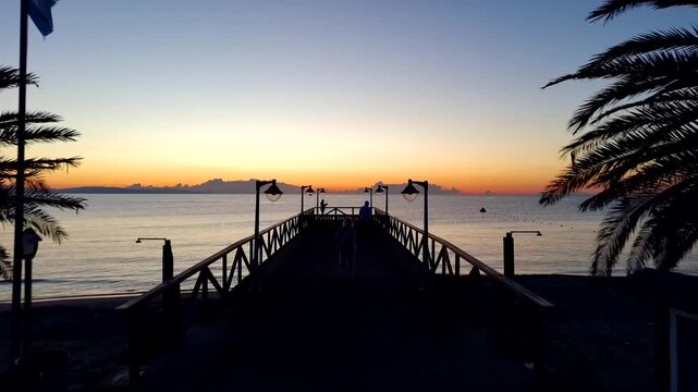 Aerial view of a wooden pier extending into the sea at sunrise with palm trees and people walking on the deck in Paralia, Katerini, Greece.