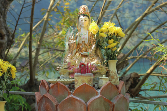 Outdoor Buddhist altar with a golden Lady Buddha Guanyin statue and yellow chrysanthemum flowers by the river in Phong Nha, Vietnam.