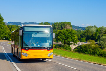 Yellow regional passenger bus driving along a scenic countryside asphalt road under a bright blue sky surrounded by green trees and hills © Sergio Grisaldi