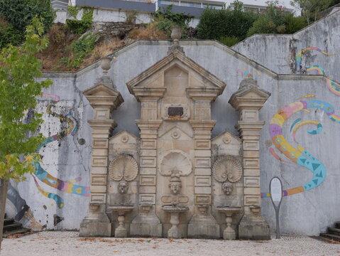 View of the historic stone fountain Fonte das tres bicas with its ornate carvings and shell motifs, set against a wall with colorful murals in Covilha, Castelo Branco, Portugal.