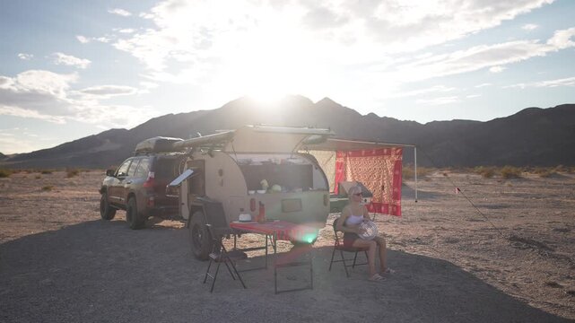 A mature woman enjoys playing a doumbek drum next to her teardrop trailer while overlanding in the Mojave desert at sunset.