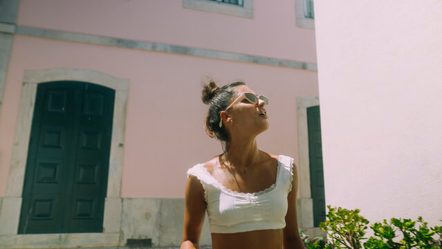 Young woman in white sleeveless top and sunglasses gazes upward beside pastel pink Mediterranean building with stone window frames and dark green doors on sunny day