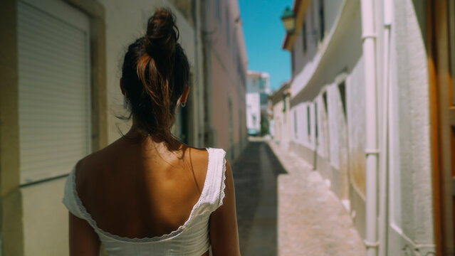 Young woman with hair bun walking away on sunlit narrow Mediterranean street from rear view