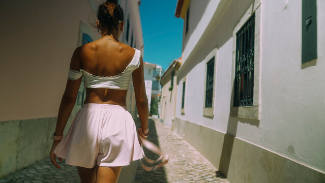 Rear view of young woman walking down sunlit narrow Mediterranean street, white off-shoulder crop top and pale skirt, holding sandals, blue sky and whitewashed buildings