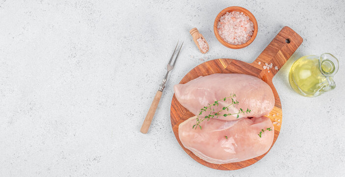 Raw chicken breast fillets with herbs on wooden cutting board, kitchen knife, salt bowl, pepper grinder, and oil bottle in top view food preparation scene