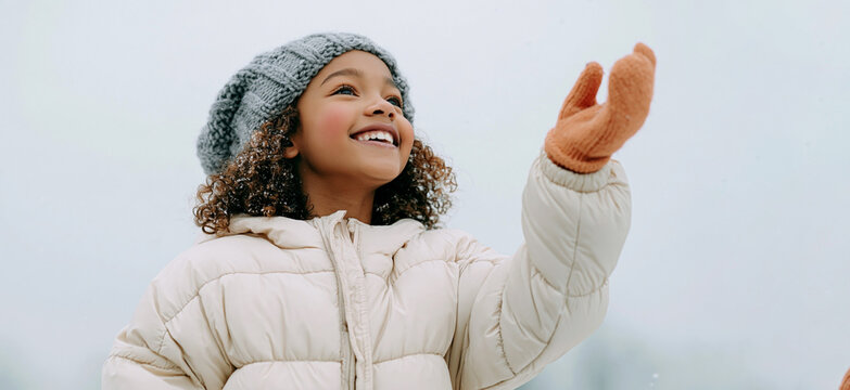 Smiling child in winter coat and knitted hat reaching toward falling snow against a pale outdoor sky, joyful seasonal portrait with warm mittens and curly hair