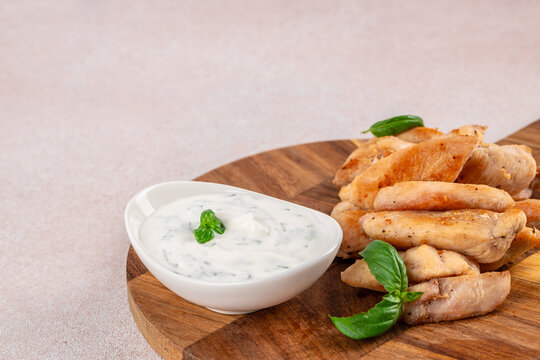 Golden fried chicken strips with creamy herb dipping sauce and fresh basil on a round wooden serving board, appetizing food still life