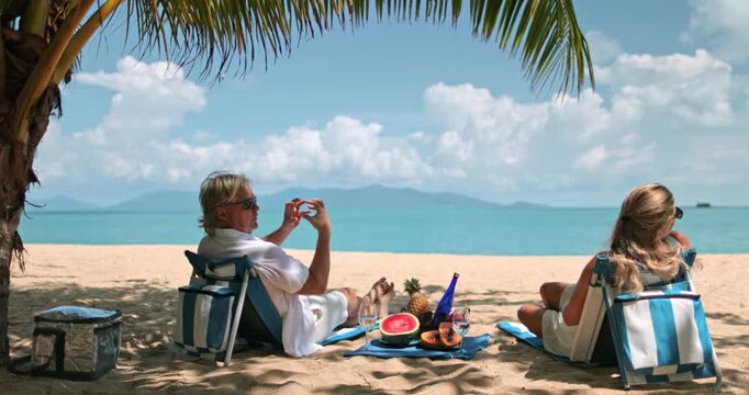 Tropical vacation on Samui Island, Thailand. Mature Caucasian couple relaxing on beach chairs under a palm tree, having a picnic with fresh fruit and drinks while enjoying the beautiful ocean view