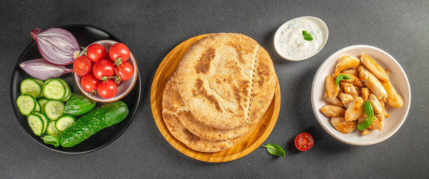 Flat lay of pita bread with fresh vegetables and grilled dumplings arranged on plates over a dark tabletop, Mediterranean inspired food composition