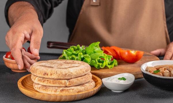 Flat lay of stacked pita breads with fresh lettuce, tomato, dipping sauce, and cook preparing ingredients on a wooden board in a kitchen setting