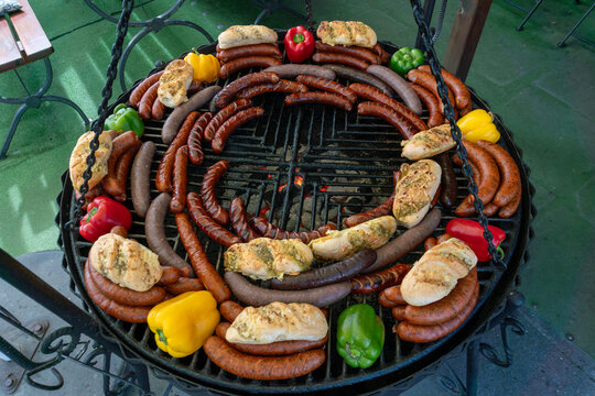 Large round charcoal barbecue grill loaded with assorted sausages, bread rolls and red, yellow and green bell peppers. Glowing coals and grilled food create a rich outdoor catering scene ideal for bar