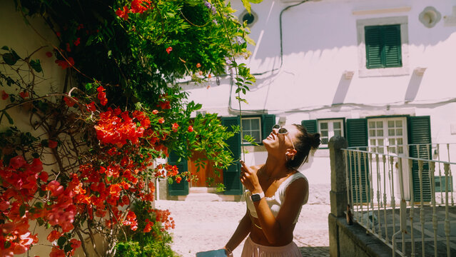 Smiling young woman in sunglasses reaching toward red bougainvillea on Mediterranean street beside white stucco wall, green shutters and balcony railing in warm sunlight