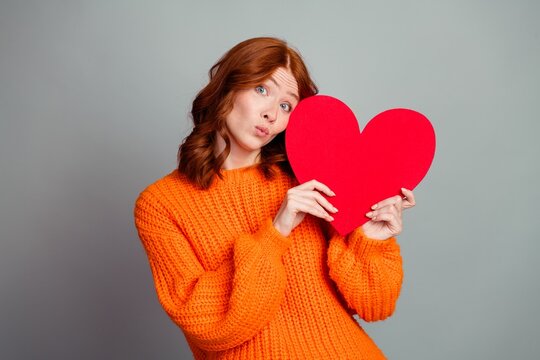 Young redhead woman in orange sweater holding a red heart and making a playful kissy face, portrait expressing love, affection and Valentine mood for romantic greeting or social media