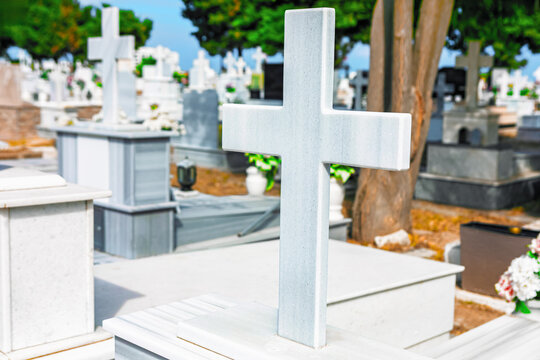 Marble cross marks a grave in a cemetery, with marble monuments and headstones arranged among trees in the background. Flowers and grave ornaments sit atop the surrounding tombs