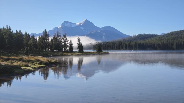 Maligne Lake reflects snow-capped mountains and green evergreen trees under a clear blue sky, with mist rising from the calm water in Jasper National Park, Alberta, Canada