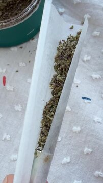 Close overhead shot of fingers filling and pressing shredded dried herb into rolling paper on tufted fabric, with open metal container visible at upper left corner