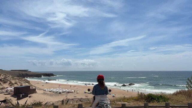 Rear view helmeted scooter rider pausing at coastal overlook with sandy beach rocky headland and wide blue sky
