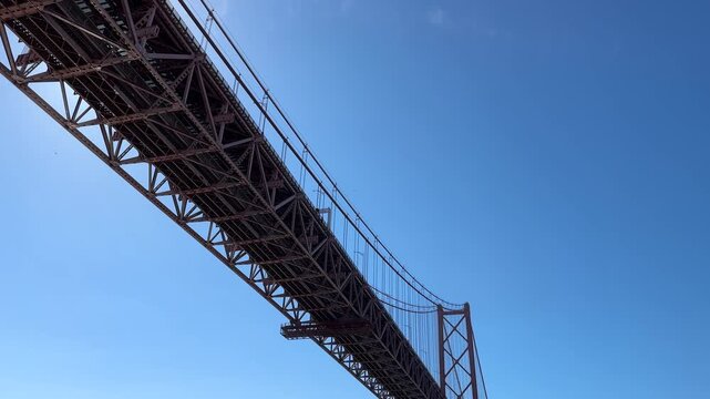 Low angle view of 25 de Abril Bridge from Doca de Santo Amaro waterfront in Lisbon Portugal, dramatic steel suspension structure crossing the Tagus River under a clear sunny blue sky