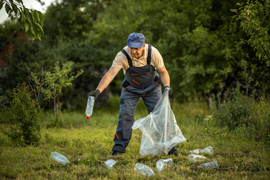 Worker picks up plastic bottle beside open trash bag in grassy, tree lined area. Focused man gathers scattered litter, supporting environmental cleanup and responsible waste disposal.