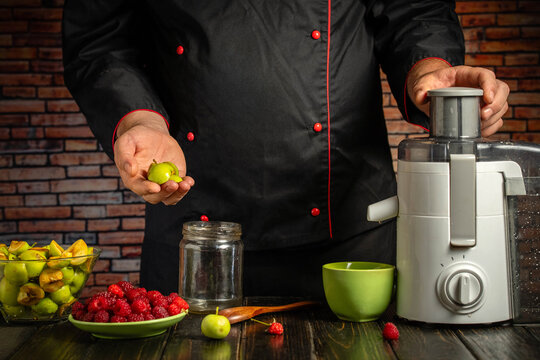 Cook presents green apples beside countertop juicer, with raspberries and sliced fruit arranged nearby. Kitchen equipment and glass jar suggest fresh juice preparation in brick walled setting.