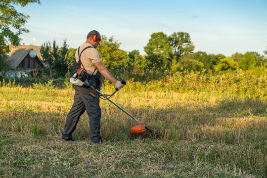 Man trimming field grass with brush cutter in rural meadow. Worker wears hearing protection and safety gloves, standing in bright evening light while managing overgrown vegetation near farmhouse.