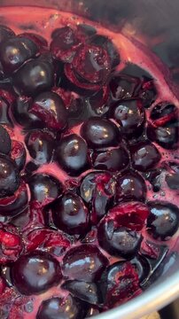 Close-up of halved cherries simmering in metal pot, glossy deep purple juice bubbling with bright magenta foam, showing split skins and tender fruit flesh for recipe preparation
