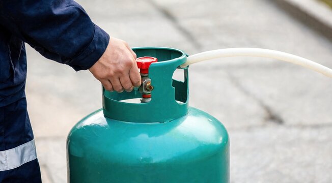 A person in a dark blue jacket holds a teal propane tank with a red valve and white hose attached, positioned outdoors on a concrete surface.