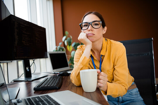 Bored female programmer at office desk holding coffee and staring at computer screens during a long workday