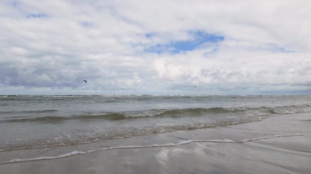 Waves breaking on sandy beach with cloudy sky and distant kite surfers