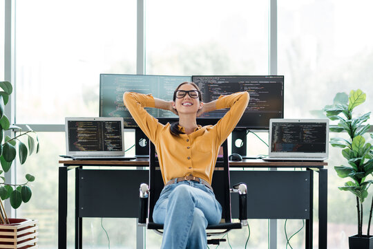 Relaxed female programmer leaning back at her desk with multiple monitors showing code in a bright modern home office