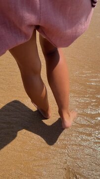 Rear low-angle view of legs and pink skirt walking along sunlit sandy shoreline, stepping through wet sand and leaving clear footprints in warm golden sunlight