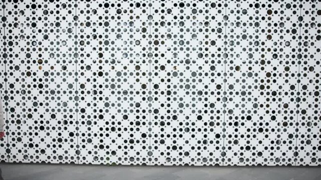 Man walking past perforated white metal wall with repeating circular pattern on urban street sidewalk as shadows shift across facade while wearing light jacket and glasses