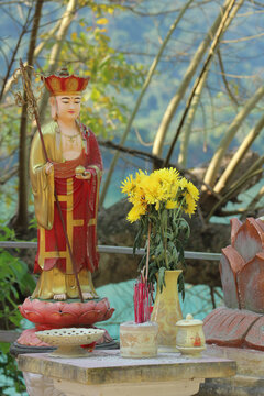 Outdoor Buddhist altar with a colorful statue of Ksitigarbha Bodhisattva or monk holding a staff next to yellow flowers in Phong Nha, Vietnam.