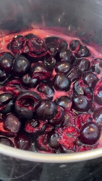 Overhead view of cherries simmering in stainless pot as dark skins split, releasing deep purple-red juice, bubbling into glossy syrup with pink foam