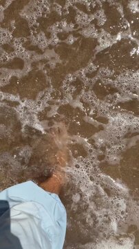 Overhead barefoot step on wet golden beach sand at shoreline, sunlit shallow surf and foamy water with first-person viewpoint of bare feet walking forward