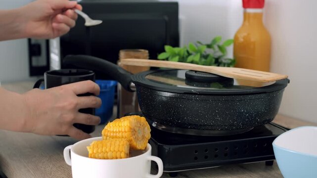 Hungry person mixes beverage with spoon next to corn on cob bowl, managing malnutrition risk during meal prep. Individual cooks food using compact electric stove in simple kitchen area.