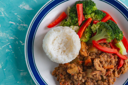Freshly cooked lentils, rice, and vegetables in a white bowl. Bright red peppers contrast with green broccoli. Blue rimmed plate holds healthy meal. Texture of grains and veggies is crisp and inviting