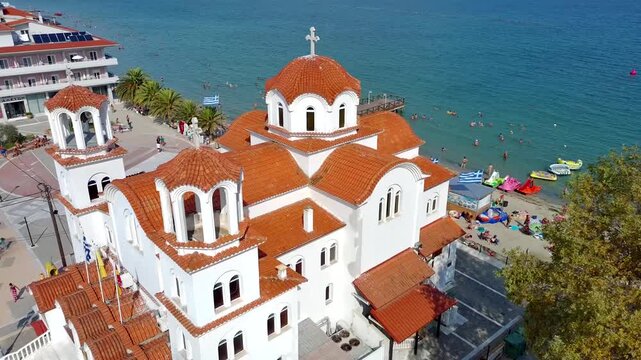 Aerial view of the white Agia Fotini Church with orange terracotta roofs situated by the sandy beach and turquoise sea in Paralia Katerinis, Pieria, Greece.