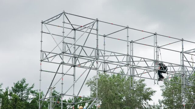 Outdoor galvanized scaffold structure with construction workers standing on upper platform under overcast sky, diagonal braces and safety tape visible across steel grid