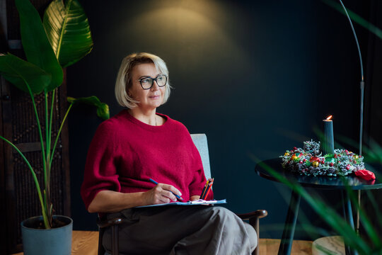 Middle aged woman planning goals for next new year while sitting on armchair in cozy dark home interior. Self-reflection concept. Personal Year review. Coaching tools. Finding Balance in Life.