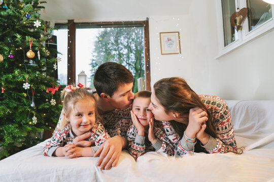 Happy family in festive pajamas hugging on bed on Christmas Eve in decorated apartment. Dad, mom,daughter, son are lying on bed and having fun together. Family celebrating winter holidays at home