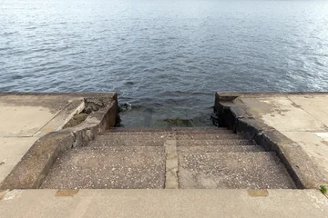 Worn concrete steps descend into calm rippling water at a weathered waterfront pier, highlighting texture, erosion and quiet atmosphere. Ideal for coastal, urban decay, travel or landscape concepts © evannovostro