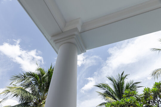 A white architectural column supports a covered porch framed by lush palm trees and bright blue sky, evoking tropical resort style, relaxation, coastal living and sunny outdoor architecture