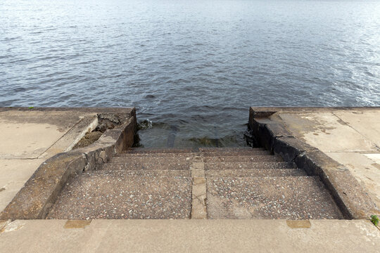 Worn concrete steps descend into calm rippling water at a weathered waterfront pier, highlighting texture, erosion and quiet atmosphere. Ideal for coastal, urban decay, travel or landscape concepts