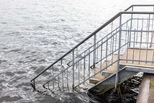 A rusted metal staircase with railings descends from a pier into rippling river water, partially submerged and weathered, evoking a moody, neglected riverside or industrial waterfront scene