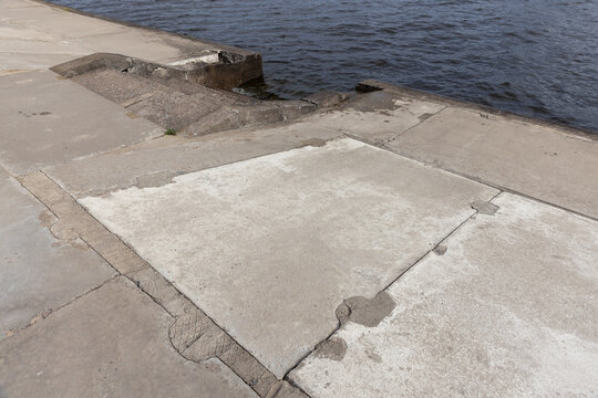 Weathered concrete slabs form a cracked waterfront pier edge beside calm water, showing texture, erosion and urban coastal decay. Ideal for backgrounds, construction and industrial concepts