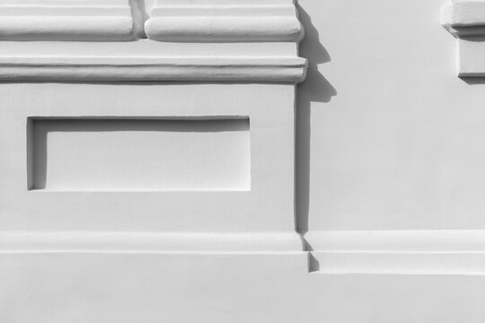 Minimalist white stucco facade featuring classical molding and rectangular reliefs. Sharp shadows emphasize texture, lines and geometry, creating a clean architectural detail ideal for background