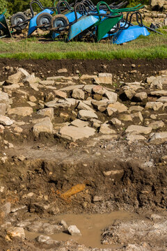 Excavation trench showing exposed stone paving and layered soil strata, row of wheelbarrows tipped above the grass bank at archaeological dig