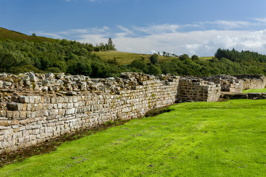 Ancient Roman masonry wall showing rough stone courses and mortar, green turf in foreground and forested slope rising in background under summer sun